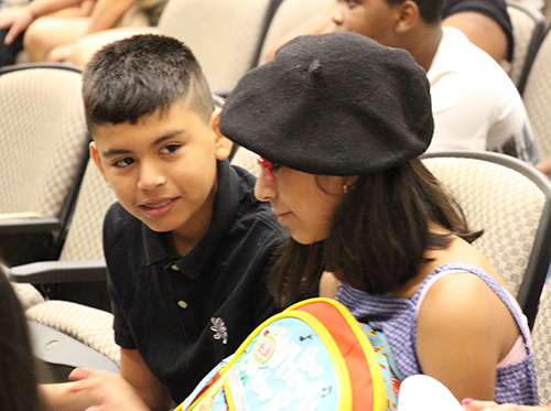 Two students, a boy on the left and girl on the right., are seated in an auditorium. The girl has shoulder length dark hair and is wearing a black beret. The boy is looking at her, while she is looking at a paper.