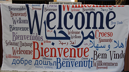 A large sign with a white background that says Welcome very large in blue. Around it in different shades of blue and red says welcome in different languages.