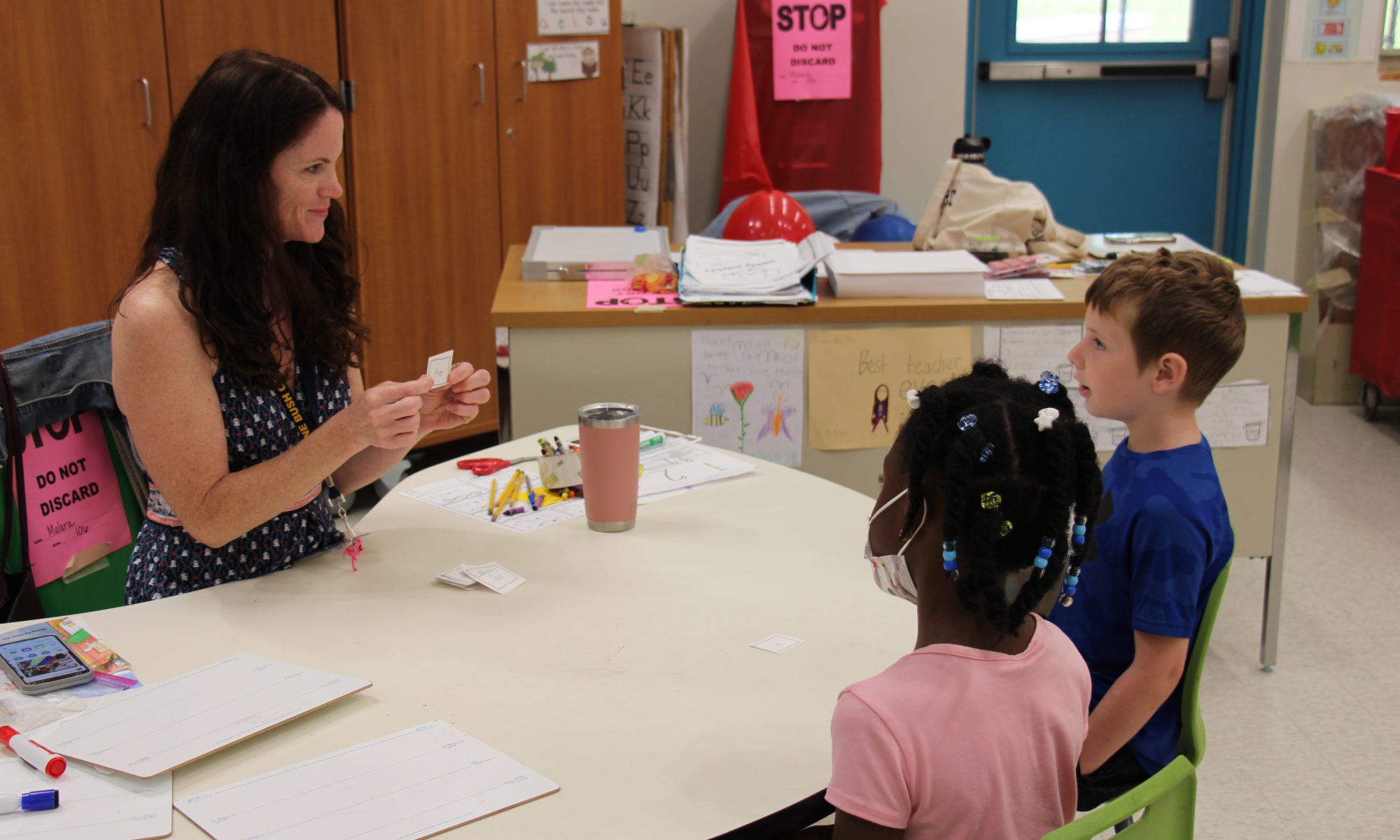A woman with long dark hair holds up a piece of paper. She is sitting at a table with two young students, a boy wearing a blue shirt and a girl wearing a pink shirt. They are looking at the teacher.