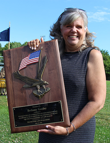 A woman with dark hair smiles as she holds a large plaque with an American flag and an eagle on it. The plaque says "Presented ot Bethany Ganley in recognition and appreciation fo ryour inspiration and leadership to the students of Pine Bush High School and the Pin eBush Leadership and Law Academy. Thanks for a job well done! July 15, 2022"