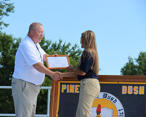 A young woman with long blonde hair, wearing a navy blue shirt and khaki pants shakes hands with a man wearing a white shirt and khaki pants. He is presenting her with a plaque.