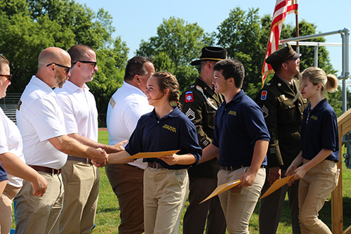 A line of adults wearing white shirts and khaki pants shake hands as students, wearing navy blue shirts and khaki pants come by.