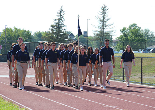 A group of about 25 high school students, all wearing navy blue shirts and khaki pants march onto the high school track. The young woman in front is carrying a platoon flag.