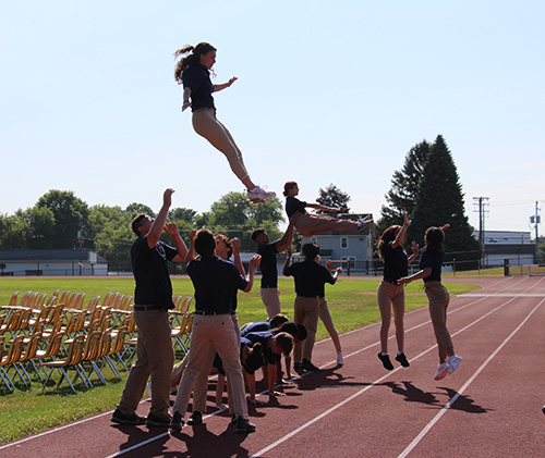 A group of high school students, all wearing navy blue shirts and khaki pants, are on a high school track. Several are looking up with their arms out as they toss a fellow student up in the air.