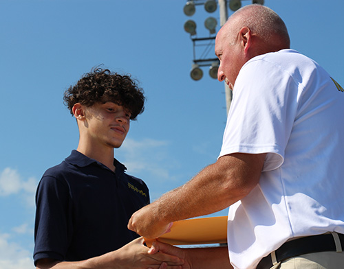 A young man with short curly hair, wearing a blue shirt, accepts a large envelope from a man wearing a white shirt. In the background is a blue sky and stadium lights.