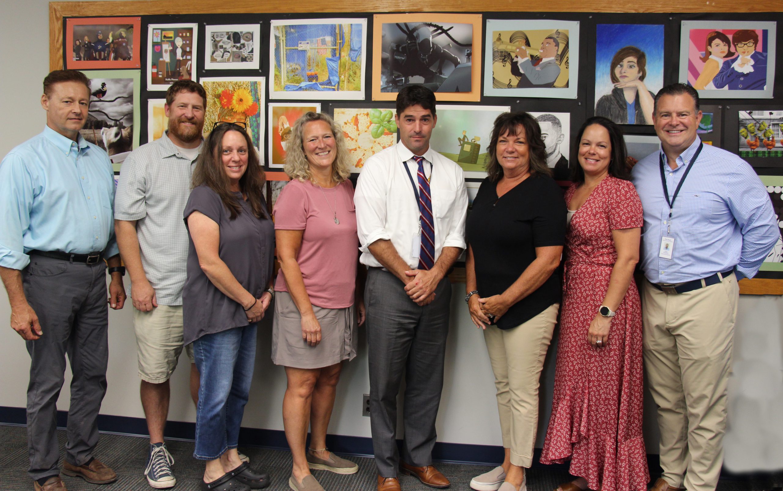 Eight adults stand in front of a wall of art projects. Two men on the left, then two women, another man, two women and a man on the right.