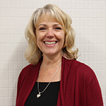 A woman with shoulder length blonde hair smiles. She is wearing a black dress and maroon sweater with a silver necklace. The background is a white tile wall.