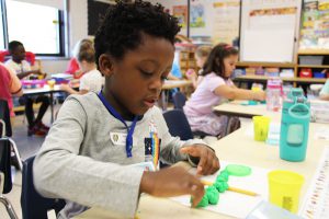 A young elementary student with black hair wearing a gray shirt sits at a table playing with green playdoh. There are other children in the background playing too.