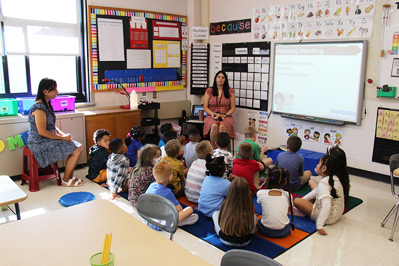 A group of younger elementary students sit on a rug as a teacher, who is sitting in front of them, talks to them. There is another woman sitting behind them.