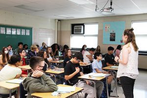 A woman with semi-long dark hair stands in front of a classroom. She is wearing a white shirt and black pants. She is talking to a group of about 25 middle school kids who are all sitting at desks.