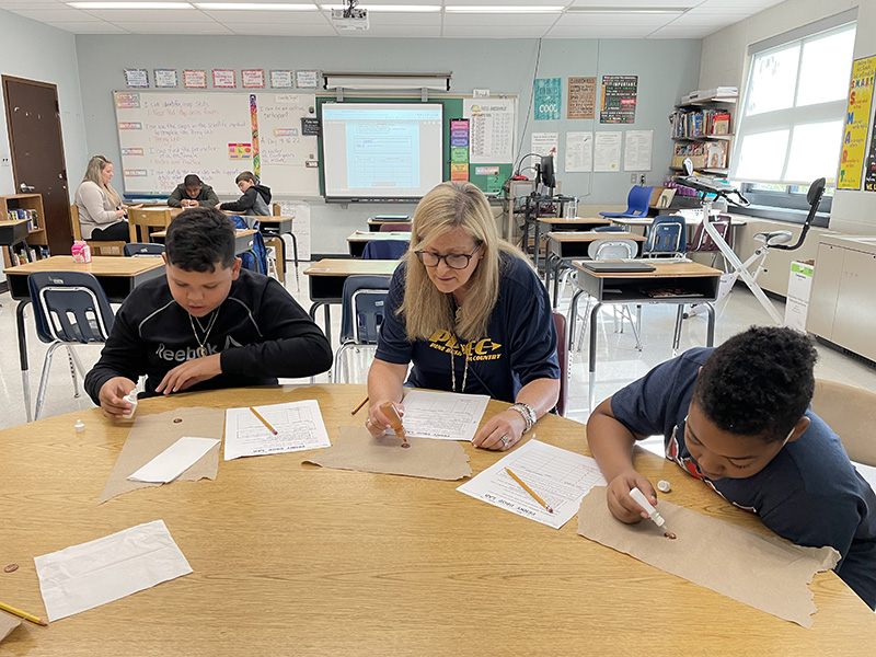 Two sixth grade boys sit at a table putting drops of water on a penny. In between them is a woman with long blonde hair doing the same. They are in a classroom.