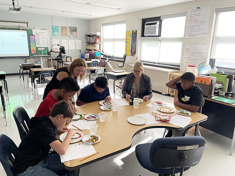 A group of sixth grade students - four of them sitting at a table working on an experiment. There is a woman at the table working with them and another woman standing over them helping.