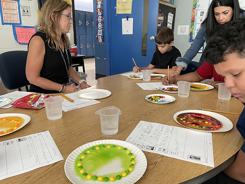A woman with long blonde hair sits at a table helping students do an experiment. There is a plate with green and yellow candies on them with water making a pattern. There is another woman leaning over the table to help.