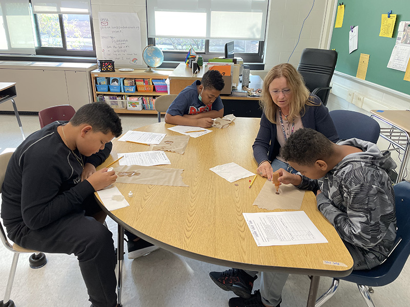 A woman with shoulder-length blonde hair works with three sixth-grade boys at a semi-circle table. They are working on a science experiment.