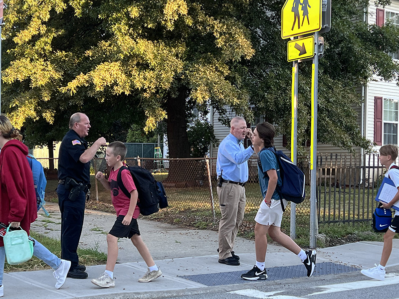 Tree in the background, sunny day. A police officer in a uniform fist bumps a kid as he walks with a backpack on. Another man with khaki pants and blue long-sleeve shirt fist bumps another kid.