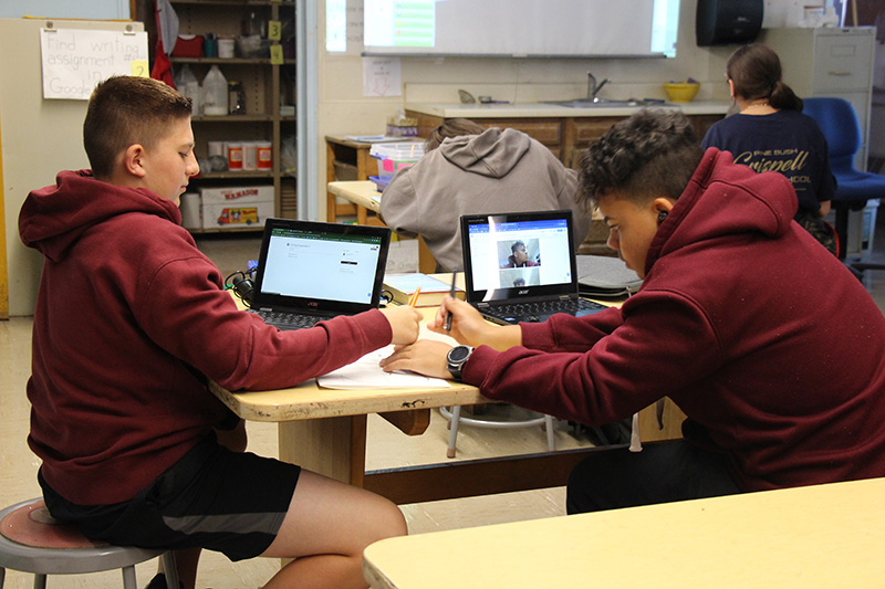 Two middle school-age young men sit at a table drawing. They both have short hair and red shirts. There are Chromebooks on the desks.