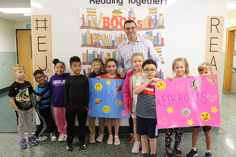 A man with dark hair and glasses smiles as he stands with a group of  10 elementary age kids. The students are holding colorful signs that say EJR Rocks.