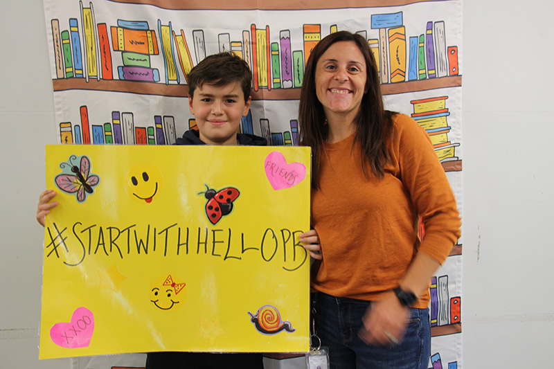 An older elementary age boy with short dark hair smiles as he holds a yellow sign that says Start with Hello PB. There are smiling faces, hearts and butterfly stickers on the poster. There is a woman with him, with long dark hair, wearing an orange shirt. She is smiling too.
