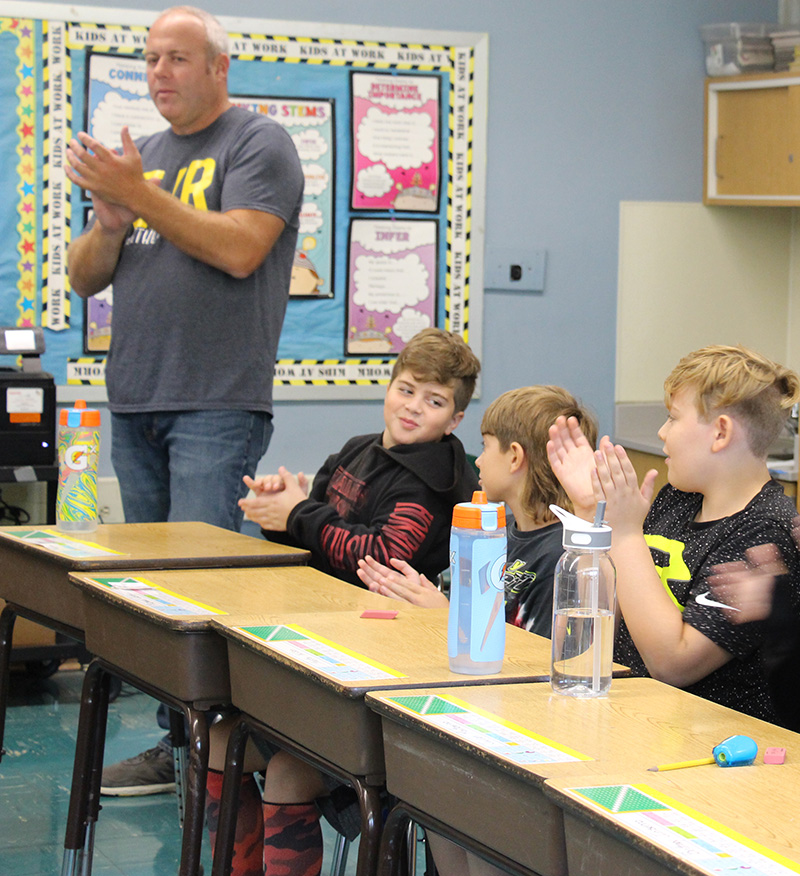 A man wearing a gray shirt that says EJR on it claps. He is standing next to a row  of desks where boys are sitting and clapping.