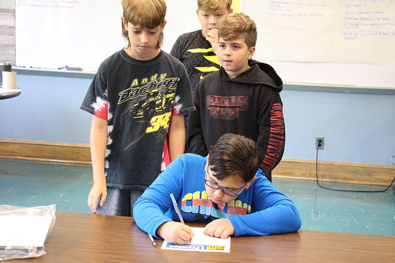 A fifth-grade boy leans over a desk and signs his name to a piece of paper. Behind him are three boys waiting to do the same.