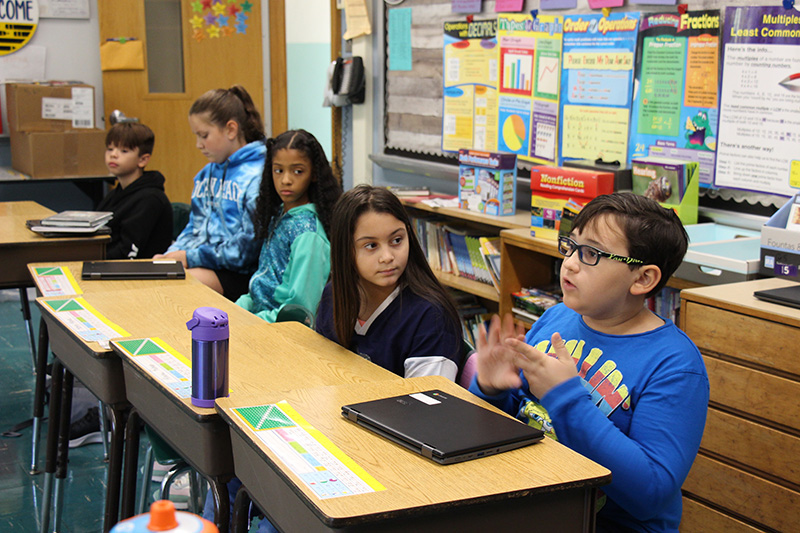 A boy wearing glasses and a long-sleeve blue shirt sits at his desk and talks using his hands. On his desk is a chromebook and next to him are four students who are listening. The girl next to him has long dark hair and is watching him intently.
