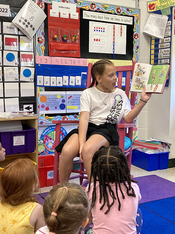 A third grade girl, wearing a white shirt and dark shorts, with her hair back in two braids, sits on a chair in a classroom holding up an open book. She is reading to kindergarten students who are sitting in front of her on the floor listening.