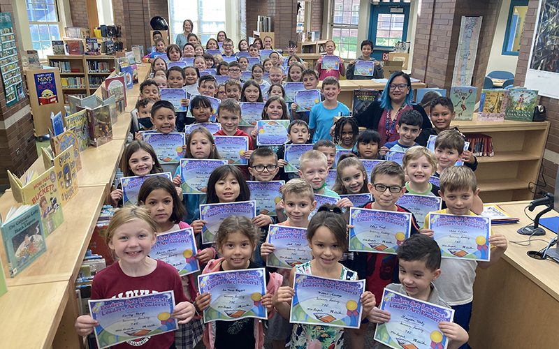 A sea of elementary children - about 65 - all holding blue certificates for the reading they did in the summer. There is an adult way in the back and another on the right. They are in a window-filled library.