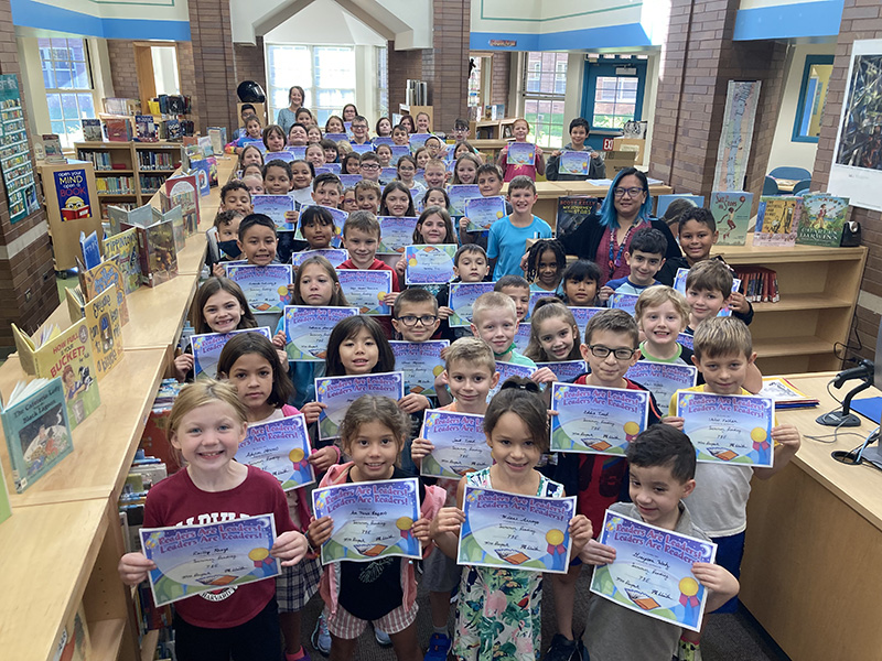A sea of elementary children - about 65 - all holding blue certificates for the reading they did in the summer. There is an adult way in the back and another on the right. They are in a window-filled library.