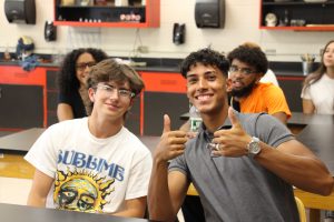 Two young men sit at a table in a classroom and smile. the kid on the left has longer short brown hair, is wearing glasses and a white tshirt that says Sublime in blue on it with a drawing of a creepy face underneath. The boy on the right is giving two thumbs up. He is wearing a gray polo shirt, has short dark hair and is wearing a watch. There are students behind them in the classroom.