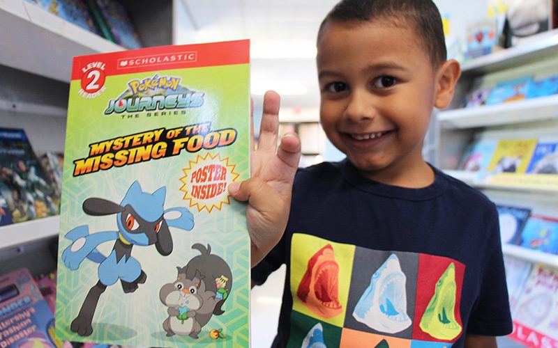 A kindergarten boy with very short dark hair smiles as he holds up a paperback Pokemon book.