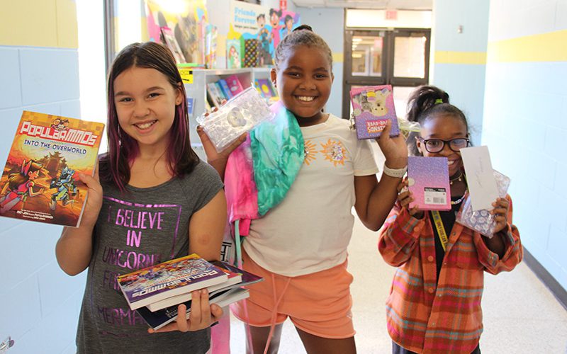 Three older elementary girls stand together smiling holding up several books each. they are all smiling.