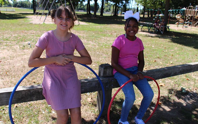 Two girls sit on a wooden fence. The girl on the left has her dark hair in ponytails. She is wearing a pink dress with short sleeves and is leaning on a blue hula hoop. The girl on the right is wearing a pink shirt, blue jeans and sneakers. She has a big white bow in her hair and a small braid with white beads hanging down on the side of her face. Both girls are smiling. There is grass and a swingset and other playground equipment in the background.