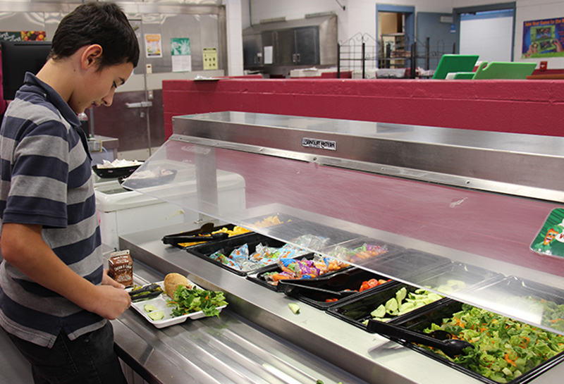 An older elementary student with short dark hair wearing a blue and gray striped polo shirt puts salad on his lunch tray, which also has a roll and chocolate milk.