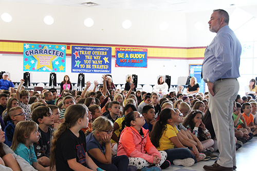 A man wearing light colored pants and a blue shirt stands in front of a large group of children who are all sitting on a floor.
