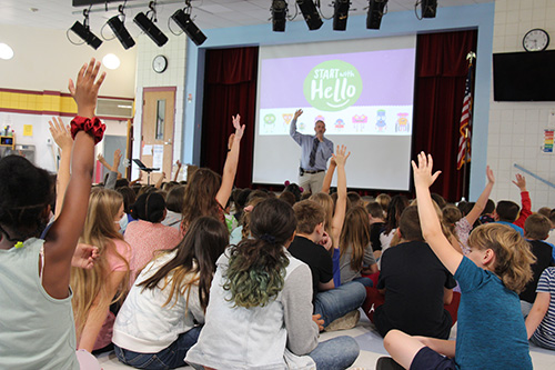A man in a blue shirt and tie stands in front of a large group of children. His hand is up in the air and many of the students are raising their hands as well. There is a screen behind him that says Start With Hello.