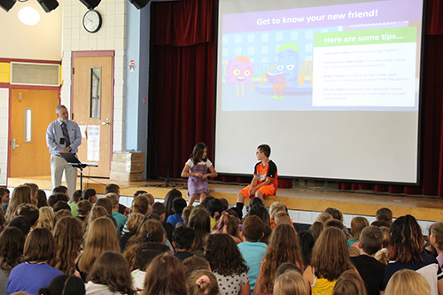 Two elementary students sit on the edgte of a stage talking to each other. There is a large group of children sitting in front of them watching and a man on the left watching them.