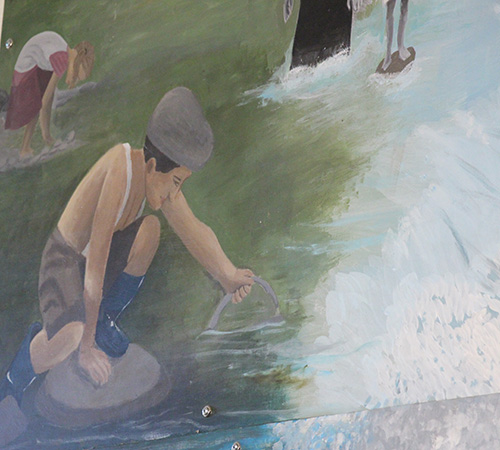 Part of a painted mural showing a little boy from the 1800s standing on a rock in the middle of a creek holding a fish. He has suspenders on and a cap.