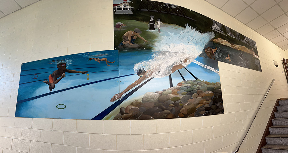 A mural, made up of four 4x8 feet pieces of masonry hang on a wall. The mural shows buildings, old and new, and kids and adults. One kid is diving into a pool.
