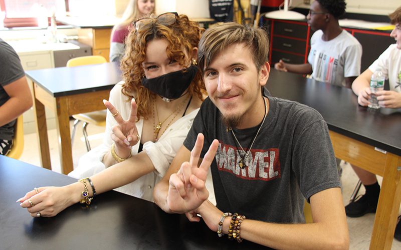 Two high school students sit at a desk and give a peace sign. The girl on the left is wearing a cream colored shirt and has necklaces on. She has curly red hair and glasses on top of her head. She is wearing a black face mask. The boy on the right is wearing a black tshirt that says Marvel in white with a red border. He has short light brown hair and a goatee on his chin. He is wearing two necklaces too. In the background are three students sitting at tables.