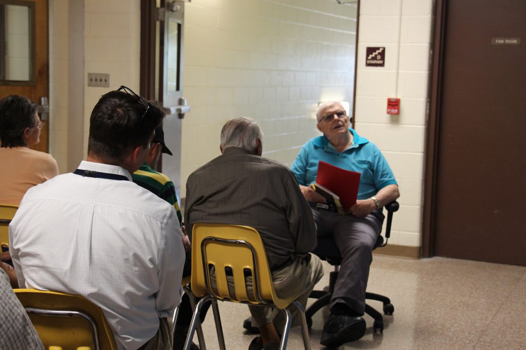 A man with white hair, wearing a blue shirt sits on a chair in front of a group of others. He is holding a folder.