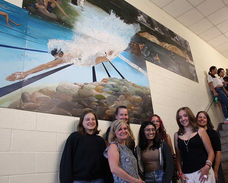 A large mural showing old buildings and kids diving into water is on a wall. Below it is a group of three adults and four young women.