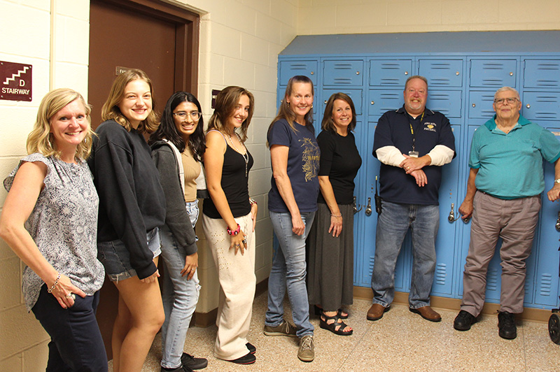 Six woman from left stand with two men on the right. There are lockers in the background.