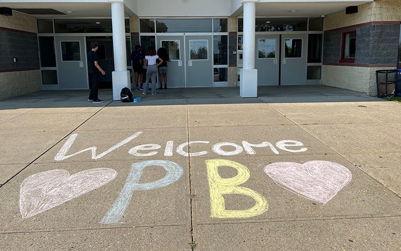 A sidewalk in front of a large school building. In chalk is written Welcome PB with pink hearts on either side of the PB. There are students up by the doors.