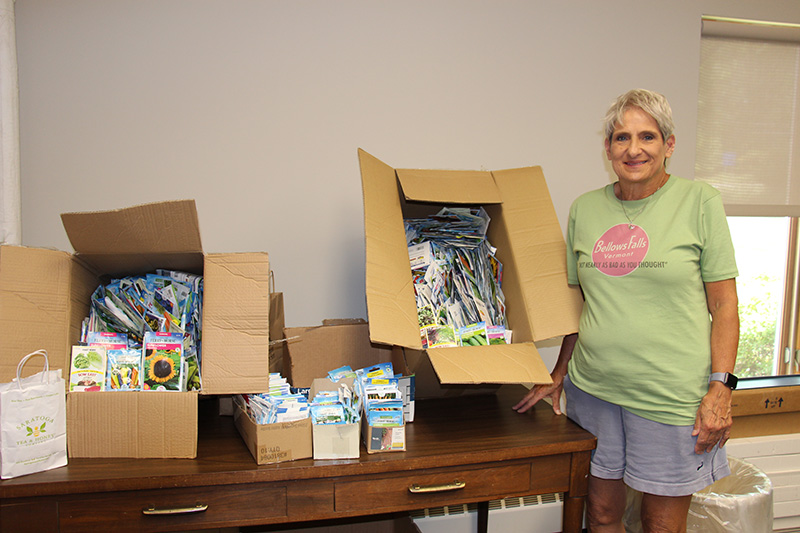 A woman with short gray hair, wearing a green shirt and gray shorts smiles. She is standing next to a table with multiple boxes, large and small, of seeds to be planted.