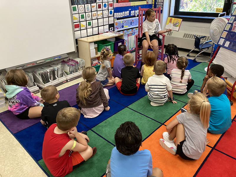 A third grade girl, wearing a white shirt and dark shorts, with her hair back in two braids, sits on a chair in a classroom holding up an open book. She is reading to kindergarten students who are sitting in front of her on the floor listening.