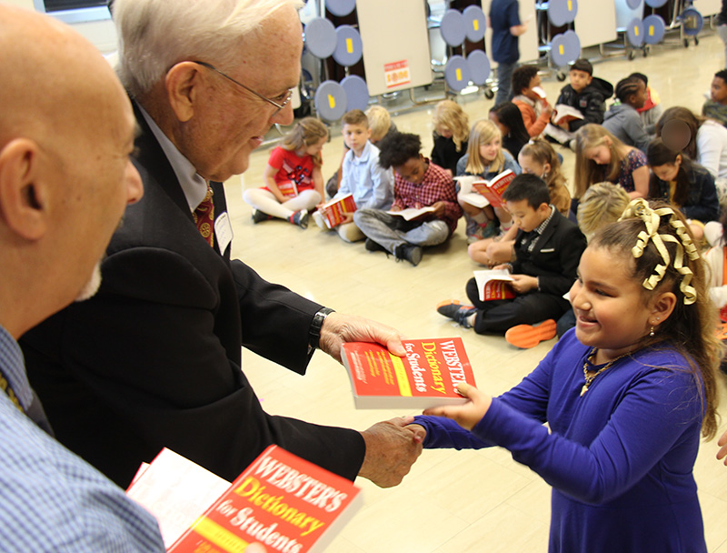 A man wearing a dark suit and glasses hands a red dictionary to a girl wearing a blue shirt. She is smiling as she shakes his hand.