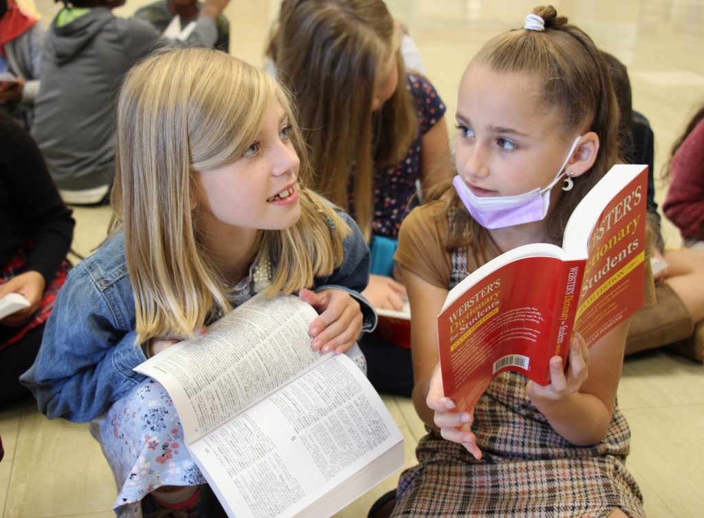 Two third-grade girls sit and look through their dictionaries. The girl on the lef5t has shoulder-length blonde hair and has her dictionary open. The girl on the right has her darker hair pulled back and is showing a page to the other girl.