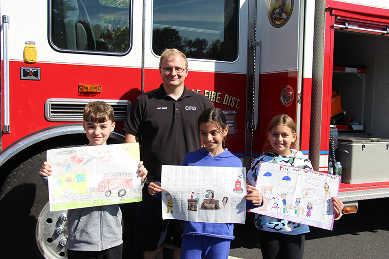 A man wearing a navy blue shirt and pants stands in front of a red and white fire truck. In front of him are three fourth grade students holding posters they made. They are smiling.