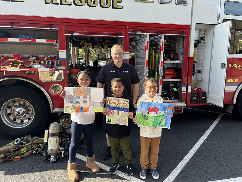 A firefighter wearing a navy blue shirt and pants stands with three third grade students. The students are holding posters they made and are smiling. There is a fire engine in the background.