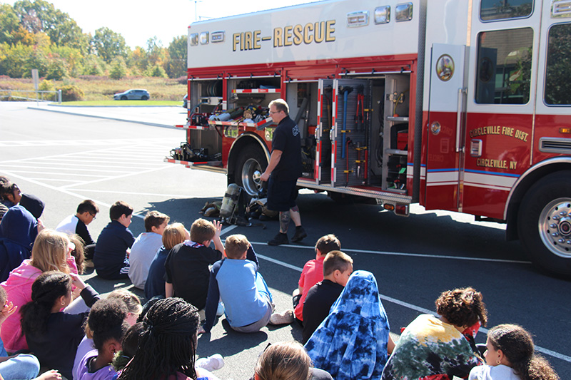 A man wearing blue shirt and pants stands in front of a red and white fire truck. He is talking to a group of elementary students sitting on the curb.